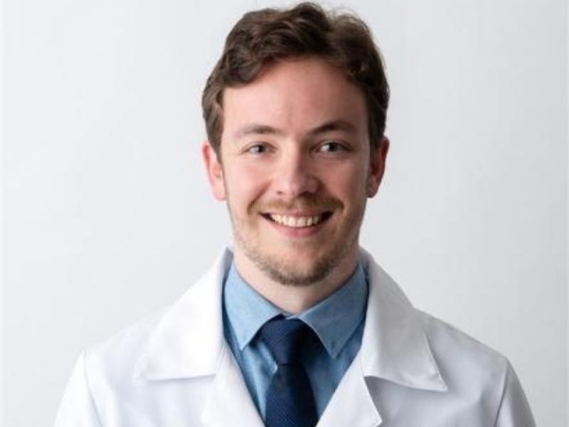 Retrato profissional do Dr. Guilherme Herrmann Matos, médico oftalmologista, terno escuro e gravata listrada, sentado em consultório oftalmológico com equipamentos de exame ao fundo, iluminação clínica realista