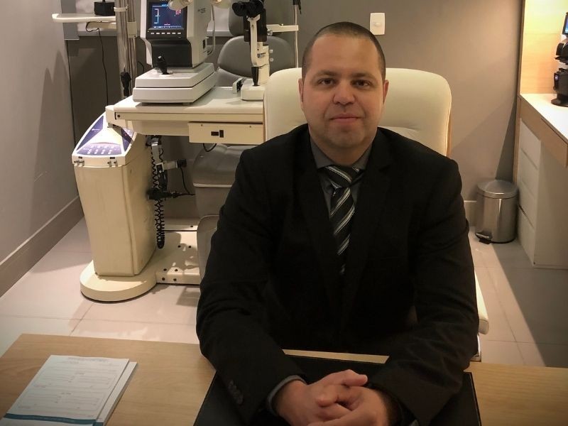 Retrato profissional do Dr. Flávio Braga da Silva, médico oftalmologista, jaleco branco, camisa azul e gravata escura, sorriso confiante, fundo neutro claro, iluminação suave de estúdio, fotografia realista